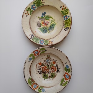 May include: Two antique, off-white ceramic plates with hand-painted floral designs. The plates feature a central floral motif in shades of orange, blue, and green, surrounded by a decorative border. The plates have a rustic, aged appearance.