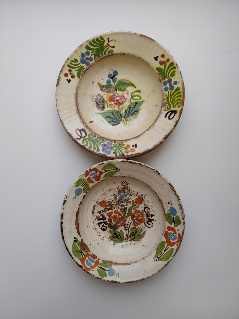 May include: Two antique, off-white ceramic plates with hand-painted floral designs. The plates feature a central floral motif in shades of orange, blue, and green, surrounded by a decorative border. The plates have a rustic, aged appearance.