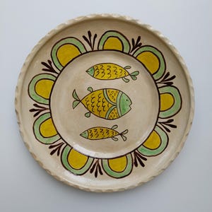 May include: A handmade ceramic plate with a cream-colored base. The plate features a decorative border of yellow and green arches, and three yellow and green fish in the center. The rim has a textured, wavy design.