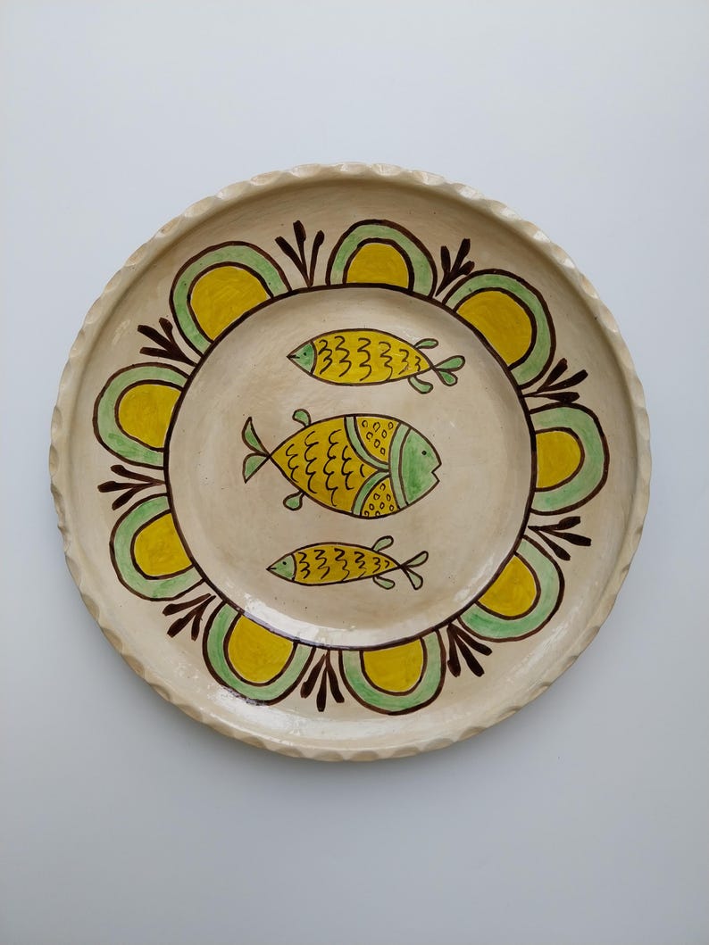May include: A handmade ceramic plate with a cream-colored base. The plate features a decorative border of yellow and green arches, and three yellow and green fish in the center. The rim has a textured, wavy design.