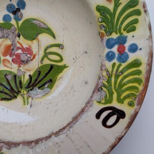 May include: Vintage ceramic plate in off-white, featuring hand-painted floral patterns in green, blue, orange, and black. The plate shows signs of wear, with cracks and chipped paint, giving it a rustic, aged look.