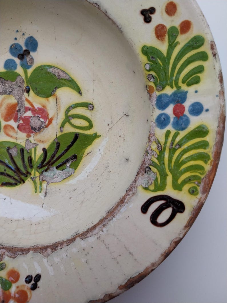 May include: Vintage ceramic plate in off-white, featuring hand-painted floral patterns in green, blue, orange, and black. The plate shows signs of wear, with cracks and chipped paint, giving it a rustic, aged look.