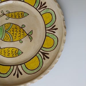 May include: A handmade ceramic plate with a cream-colored base. The plate features three yellow fish with green details and a decorative border of yellow and green arches. The rim has a scalloped edge.