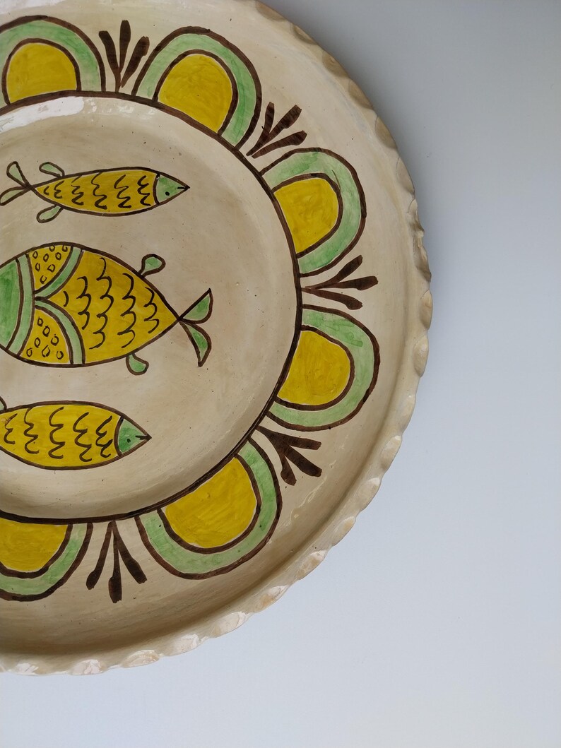 May include: A handmade ceramic plate with a cream-colored base. The plate features three yellow fish with green details and a decorative border of yellow and green arches. The rim has a scalloped edge.