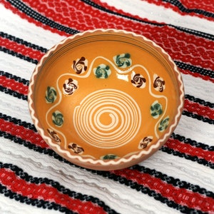 May include: A yellow ceramic bowl with a white spiral design and brown and green floral patterns. The bowl is sitting on a white and red striped fabric.