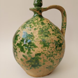 May include: A rustic ceramic jug with a bulbous body and a handle. The jug has a speckled green glaze over a tan base, with a small spout and a decorative top. The design suggests a vintage or antique aesthetic, with a handmade quality.
