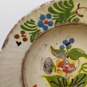 May include: A fragment of a vintage ceramic plate with a cream base and a floral design. The flowers are painted in blue, red, and orange, with green leaves. The rim has a worn, aged appearance.