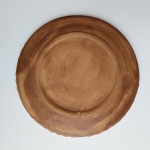 May include: A handmade, brown ceramic plate with a textured surface. The plate has a wide, slightly raised rim and a central recessed area. The underside of the plate is visible, showcasing the natural clay color and texture.