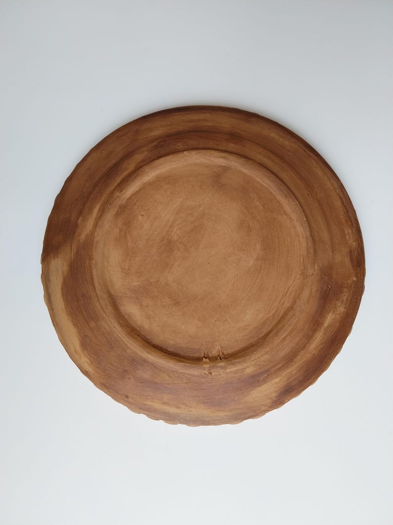 May include: A handmade, brown ceramic plate with a textured surface. The plate has a wide, slightly raised rim and a central recessed area. The underside of the plate is visible, showcasing the natural clay color and texture.