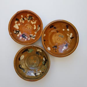 Puede incluir: Tres cuencos de cerámica hechos a mano con un aspecto rústico. Los cuencos son de color marrón cálido con detalles de pintura moteada en blanco, negro y verde. Los cuencos son redondos y tienen poca profundidad, perfectos para servir porciones pequeñas.