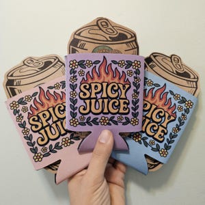 May include: Three colourful can coolers are held in a hand, featuring the text "SPICY JUICE" and "SPICY ICE". The coolers are pink, lavender, and blue, with a flame design and floral border. Cardboard can cutouts are behind.