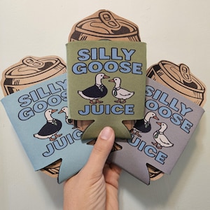 May include: Three drinks can coolers in blue, green, and grey, each with a cartoon goose wearing a suit and the words "Silly Goose Juice." Cardboard cutouts of drinks cans are behind the coolers. The coolers are held by a hand.