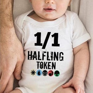 May include: A baby wearing a white onesie with the text "1/1 HALFLING TOKEN" in black. The onesie has colorful symbols below the text. The baby is lying down, held by an adult.