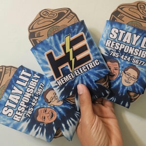 May include: A hand holding several blue tie-dye can coolers. The coolers have the text "STAY LIT RESPONSIBLY" and "HEMEL ELECTRIC" with a lightning bolt graphic. The phone number 785-424-5677 is also printed on the coolers. Cardboard can cutouts are in the background.