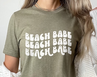 Könnte beinhalten: Ein grünes T-Shirt mit dem Text "Beach Babe", der dreimal in einer weißen, wellenförmigen Schrift wiederholt wird.