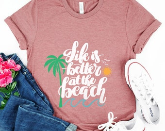 Könnte beinhalten: Ein pinkfarbenes T-Shirt mit dem Text "Life is better at the beach" in weißen Buchstaben. Das Shirt zeigt einen grünen Palmenbaum und eine gelbe Sonne.