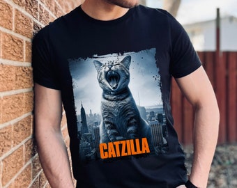 Może przedstawiać: Czarny t-shirt z grafiką przedstawiającą kota ryczącego przed panoramą miasta. Tekst "CATZILLA" jest nadrukowany na pomarańczowo pod kotem.