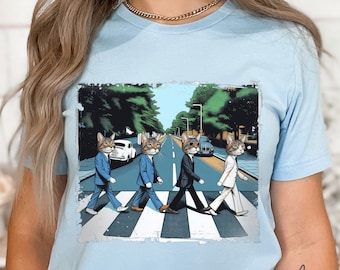 May include: A light blue t-shirt with a graphic of four cats dressed in suits walking across a crosswalk, a parody of the Beatles' Abbey Road album cover.