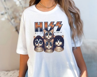 以下が含まれることがあります： KISSのメンバーに扮した4匹の猫のグラフィックがプリントされた白いTシャツ。猫の上に「HISS」という文字が黒とオレンジのグラデーションで書かれています。