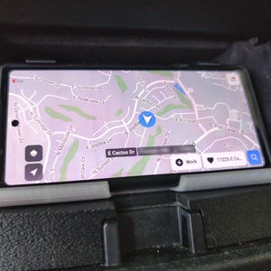 Pode incluir: Um smartphone exibe um mapa com nomes de ruas e um marcador de localização. O telefone está em um suporte preto dentro do console de um carro. O mapa mostra ruas como E Cactus Dr e E Palisades Blvd.