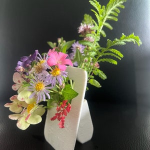 Op de afbeelding: Een witte, moderne, minimalistische vaas bevat een levendig arrangement van verse bloemen. Het boeket bevat roze, paarse, gele en rode bloemen, met groen blad. De vaas heeft een uniek, gevouwen ontwerp, tegen een donkere achtergrond.