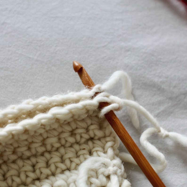 May include: Close-up of a white crocheted project with a wooden crochet hook. The hook is inserted through a loop of yarn, and the yarn is pulled through the loop.