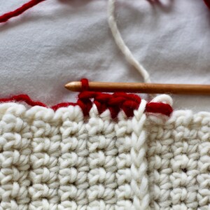 May include: Close-up of a white crocheted blanket with a red border. A wooden crochet hook is holding a red yarn strand, and a white yarn strand is visible in the background.