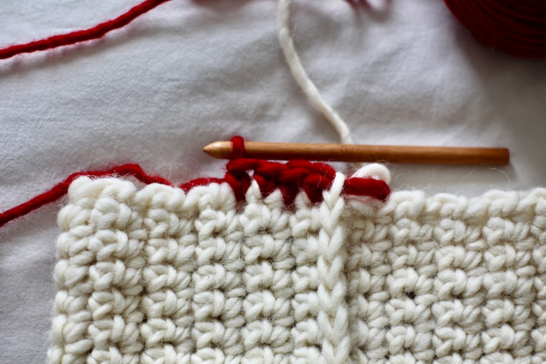 May include: Close-up of a white crocheted blanket with a red border. A wooden crochet hook is holding a red yarn strand, and a white yarn strand is visible in the background.