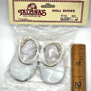 May include: A pair of white doll shoes in a plastic bag with a ruler for size reference. The shoes are made of a shiny material and have a simple design. The bag has a label that reads "Tallena's Doll Supplies, Inc. Doll Shoes Style 002".
