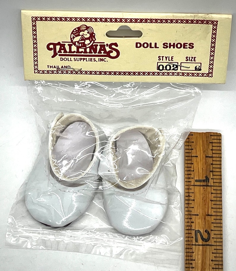 May include: A pair of white doll shoes in a plastic bag with a ruler for size reference. The shoes are made of a shiny material and have a simple design. The bag has a label that reads "Tallena's Doll Supplies, Inc. Doll Shoes Style 002".