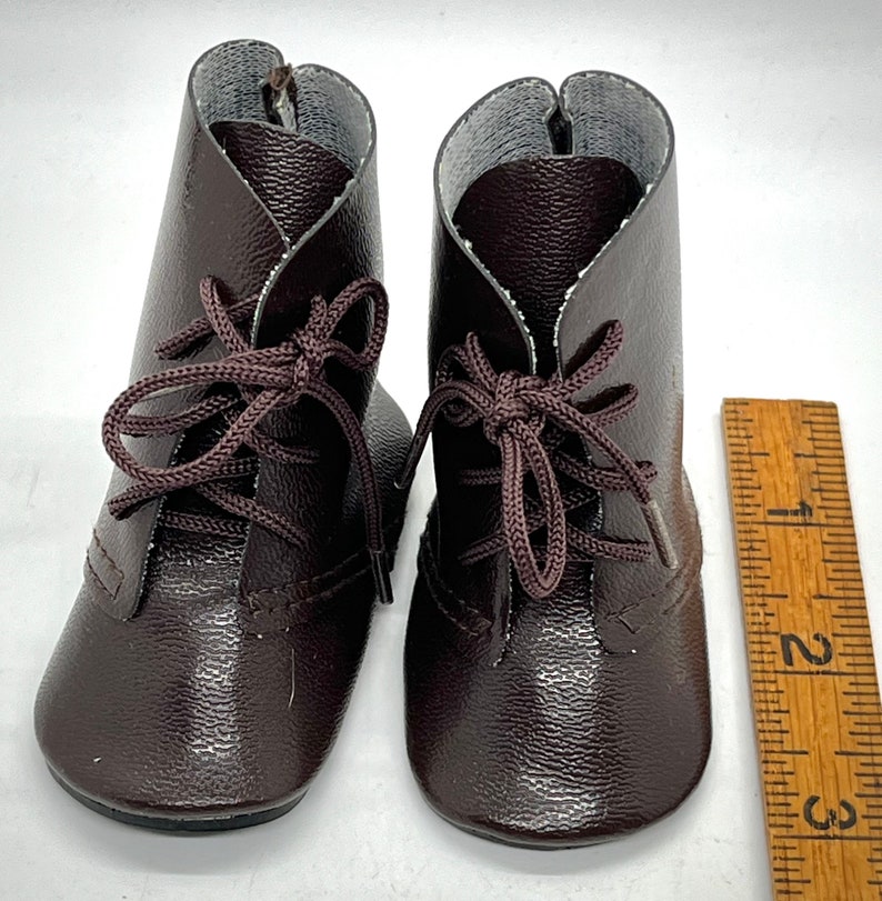 May include: A pair of brown leather lace-up boots for dolls. The boots have a textured finish and are tied with brown laces.