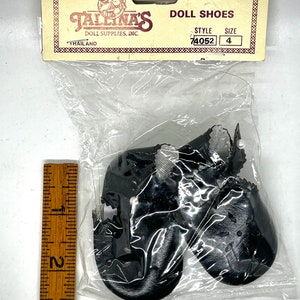 May include: A pair of black doll shoes with white trim and a pearl detail. The shoes are packaged in a clear plastic bag with a yellow and red card that reads "Jallina's Doll Supplies, Inc. Doll Shoes Style 74052 Size 4 Thailand."
