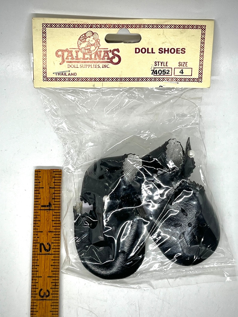 May include: A pair of black doll shoes with white trim and a pearl detail. The shoes are packaged in a clear plastic bag with a yellow and red card that reads "Jallina's Doll Supplies, Inc. Doll Shoes Style 74052 Size 4 Thailand."