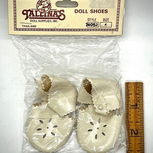 May include: A pair of white doll shoes with a pearl accent and a flower-shaped cut-out design. The shoes are packaged in a clear plastic bag with a label that reads "Tallina's Doll Shoes, Doll Supplies, Inc., Thailand, Style 74052, Size 4."