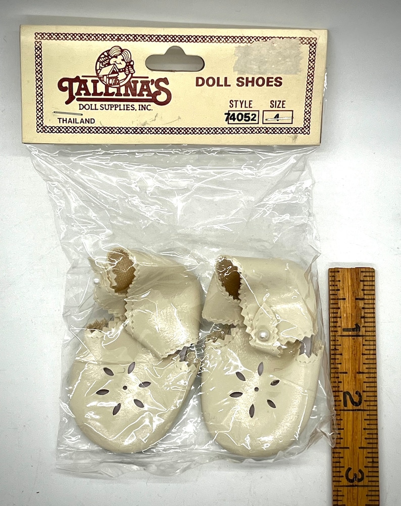 May include: A pair of white doll shoes with a pearl accent and a flower-shaped cut-out design. The shoes are packaged in a clear plastic bag with a label that reads "Tallina's Doll Shoes, Doll Supplies, Inc., Thailand, Style 74052, Size 4."