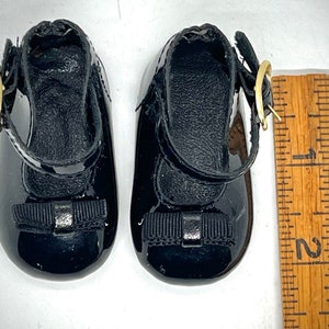 May include: A pair of black patent leather shoes with a black bow and a gold buckle. The shoes are about 2 inches long.
