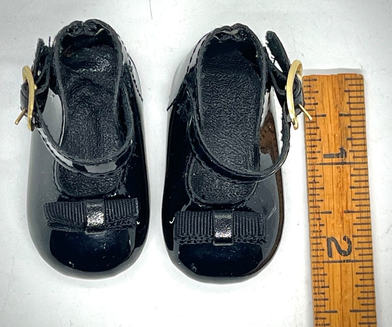 May include: A pair of black patent leather shoes with a black bow and a gold buckle. The shoes are about 2 inches long.