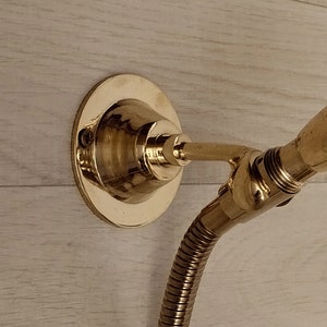 Może przedstawiać: Złota słuchawka prysznicowa z elastycznym wężem i mocowaniem ściennym. Słuchawka prysznicowa jest przymocowana do ściany za pomocą okrągłej podstawy.