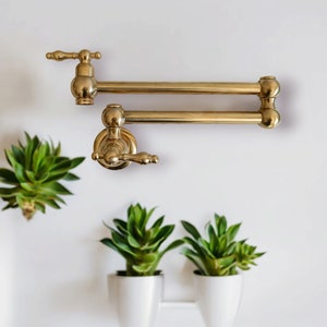 Puede incluir: Un grifo de cocina de tono dorado con un caño extensible de doble articulación. El grifo está montado en una pared blanca y tiene un diseño moderno.