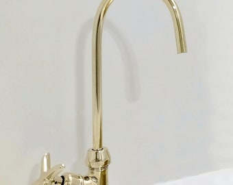May include: A gold-toned faucet with a curved spout and a cross-shaped handle. The faucet is mounted on a white countertop next to a hammered brass sink.