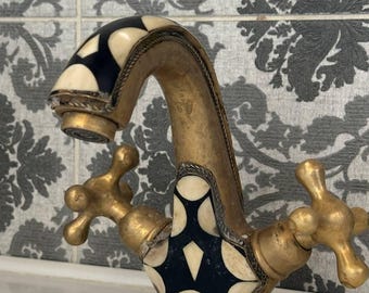 May include: Ornate brass faucet with black and cream-colored inlay. The faucet features cross-shaped handles and decorative detailing. The design includes a curved spout and is set against a patterned tile background.