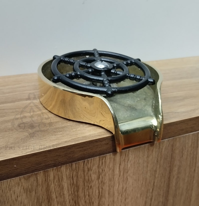 May include: A brass-coloured ashtray featuring a black ship's wheel design. The ashtray has a curved shape with a raised edge and a small lip. The ship's wheel is centrally positioned within the ashtray.