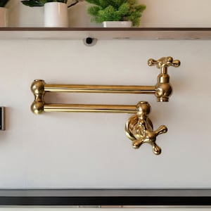 Może przedstawiać: Złoty kran kuchenny z podwójnym ramieniem i dwoma uchwytami. Kran jest zamontowany na białej ścianie.