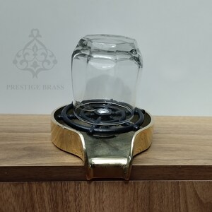 May include: A clear, faceted glass sits upside down on a brass and black metal stand. The stand has a gold-coloured rim and a black, circular interior. The text "PRESTIGE BRASS" is visible in the background.