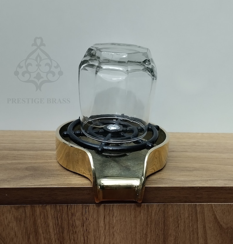 May include: A clear, faceted glass sits upside down on a brass and black metal stand. The stand has a gold-coloured rim and a black, circular interior. The text "PRESTIGE BRASS" is visible in the background.