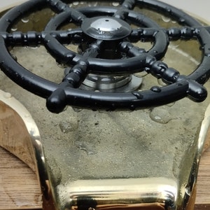 May include: A close-up of a ship's wheel-shaped faucet handle. The handle is black and sits atop a brass base. The words "PRESTIGE BRASS" are visible in the background.