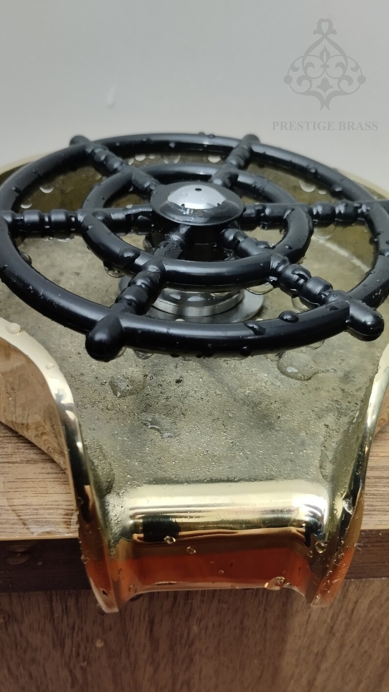 May include: A close-up of a ship's wheel-shaped faucet handle. The handle is black and sits atop a brass base. The words "PRESTIGE BRASS" are visible in the background.