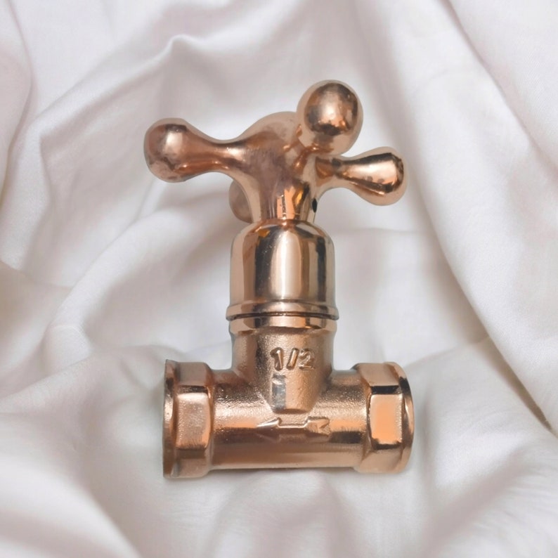 Peut inclure: Une vanne de robinet en m&eacute;tal cuivr&eacute; poli dot&eacute;e d'une poign&eacute;e en croix &agrave; quatre branches. Le corps central affiche le texte 1/2 et un symbole de fl&egrave;che indiquant le sens du flux. La vanne poss&egrave;de une base cylindrique avec des extr&eacute;mit&eacute;s hexagonales.