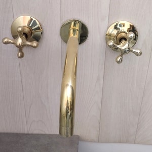 May include: A gold-toned bathroom faucet with two cross-shaped handles. The faucet has a curved spout and is mounted on a wall.