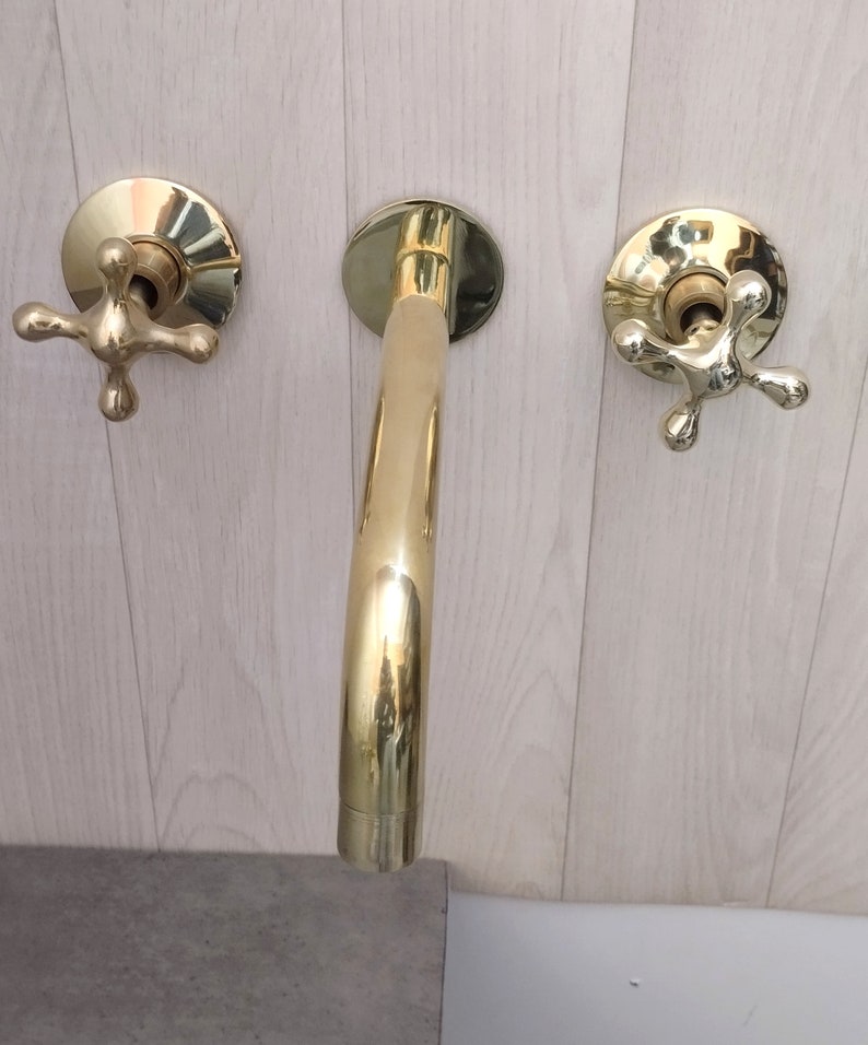 May include: A gold-toned bathroom faucet with two cross-shaped handles. The faucet has a curved spout and is mounted on a wall.